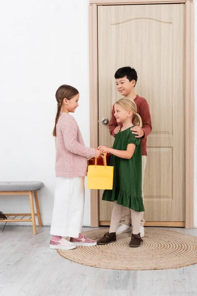 Interracial kids giving shopping bag and present to friend at home — Stock Photo