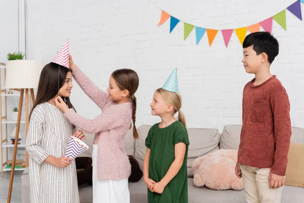 Lächelndes Mädchen mit Partymütze auf Freundin in der Nähe von interrassischen Kindern zu Hause — Stockfoto