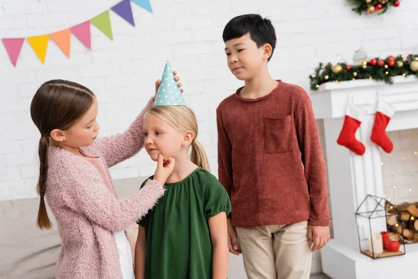 Girl wearing party cap on friend near asian boy during birthday party at home — Stock Photo
