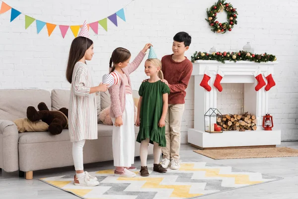 Cheerful multiethnic kids holding party caps during birthday party and winter holidays at home — Stock Photo