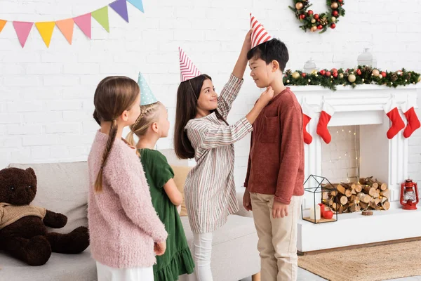 Smiling girl wearing party cap on asian friend during birthday party and christmas celebration — Stock Photo