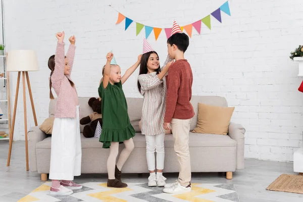 Enfants interracial joyeux dans les casquettes de fête célébrant l'anniversaire pendant la fête à la maison — Photo de stock