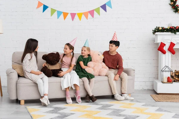 Smiling multiethnic kids in party caps sitting near soft toys and christmas decor at home — Stock Photo