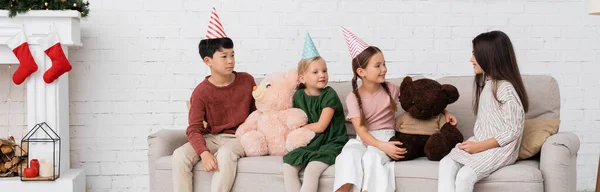 Multiethnic children in party caps celebrating birthday near soft toys and christmas decor, banner — Stock Photo