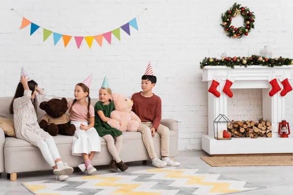 Веселые межрасовые дети в шапочках для вечеринок сидят рядом с плюшевыми мишками и рождественским декором дома — стоковое фото