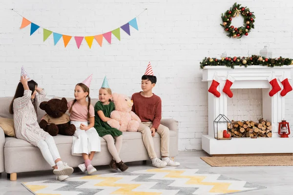 Cheerful interracial kids in party caps sitting near teddy bears and christmas decor at home — Stock Photo