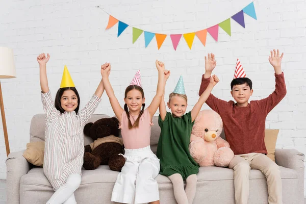 Crianças multiétnicas excitadas em bonés de festa olhando para a câmera no sofá durante a festa de aniversário em casa — Fotografia de Stock