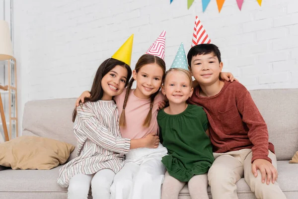Улыбающиеся межрасовые дети в шапочках на вечеринках смотрят в камеру и обнимаются во время празднования дня рождения дома — стоковое фото