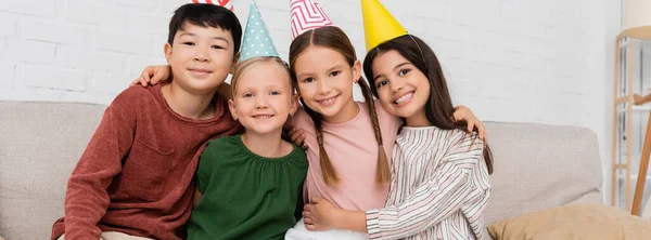 Happy interracial kids in party caps looking at camera during birthday party at home, banner — Stock Photo