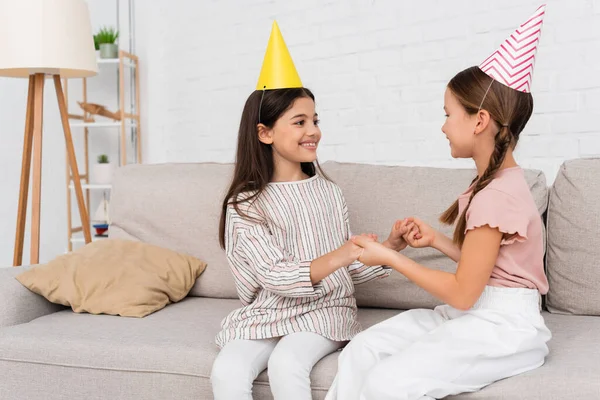 Chicas sonrientes en gorras de fiesta cogidas de la mano durante la fiesta de cumpleaños en el sofá en casa - foto de stock