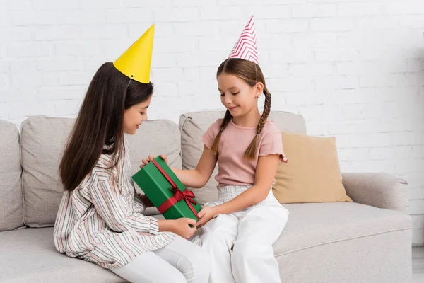 Улыбающиеся девушки в шапочках для вечеринок держат подарочную коробку с луком на диване дома — стоковое фото
