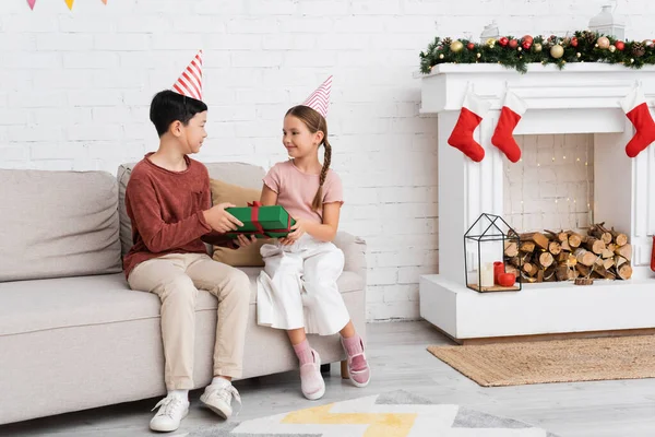 Asiático chico en partido gorra holding presente cerca amigo en sofá y Navidad decoración en casa - foto de stock