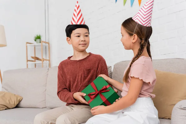 Asian boy in party cap giving present to friend during birthday party at home — Stock Photo