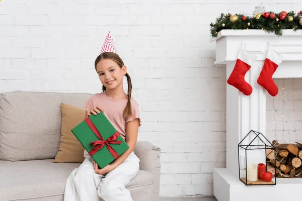Ragazza sorridente in cappello partito che tiene regalo vicino all'arredamento natalizio a casa — Foto stock