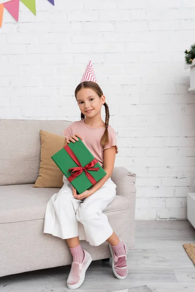 Ragazza allegra in festa tappo in possesso di scatola regalo sul divano a casa — Foto stock