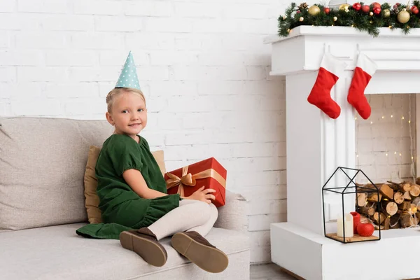 Smiling kid in party cap holding present during birthday party near christmas decor at home — Stock Photo