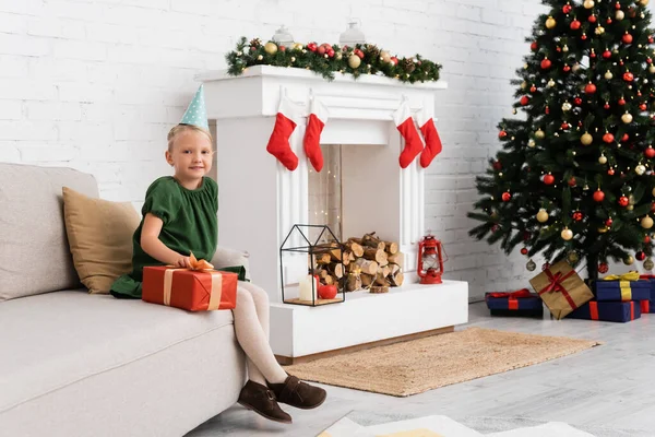 Vorschulmädchen in Partymütze hält Geschenk in der Nähe von Weihnachtsdekor und Baum zu Hause — Stockfoto