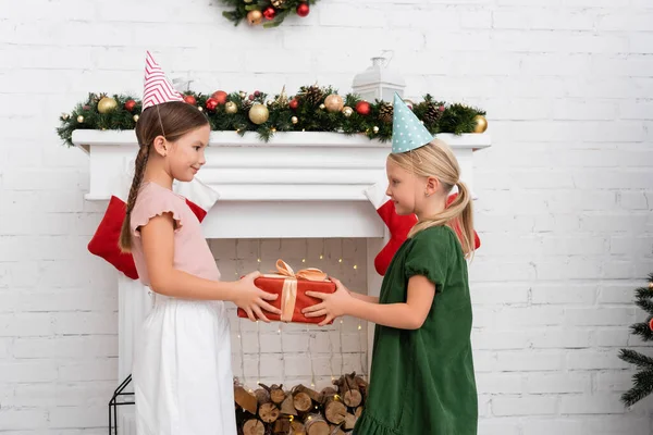 Side view of girl in party cap giving present to friend near christmas decor on fireplace at home — Stock Photo