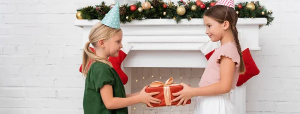 Side view of kid holding gift box near friend in party cap and christmas decor on fireplace, banner — Stock Photo