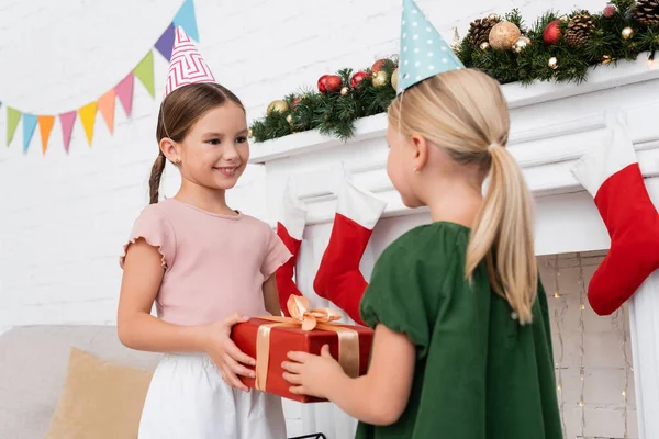 Chica sonriente en gorra de fiesta tomando regalo de amigo borroso cerca de la decoración de Navidad en casa - foto de stock