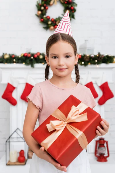 Portrait of kid in party cap holding gift box near blurred christmas decor at home — Stock Photo