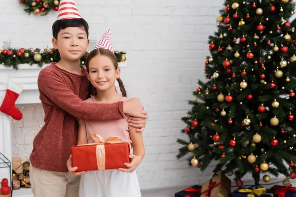 Азиатский мальчик в шапочке для вечеринок обнимает улыбающегося друга с подарком возле рождественской елки дома — Stock Photo