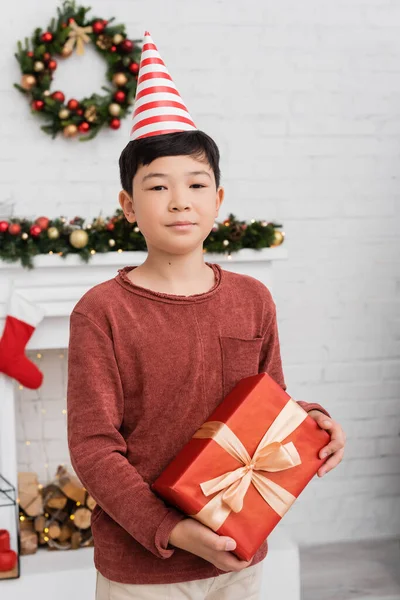 Asian boy in party cap holding present and looking at camera near blurred christmas decor at home — Stock Photo