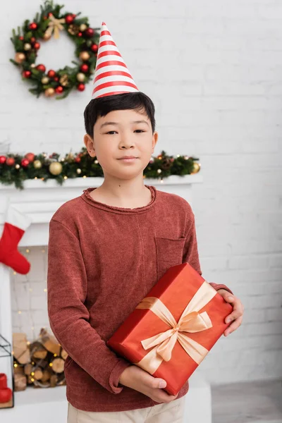 Asiatico ragazzo in partito cap tenendo presente e guardando fotocamera vicino offuscata natale arredamento a casa — Foto stock