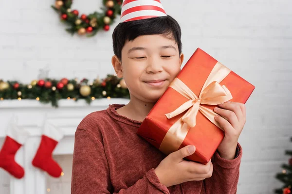 Happy asian boy in party cap holding gift near blurred christmas decor at home — Stock Photo