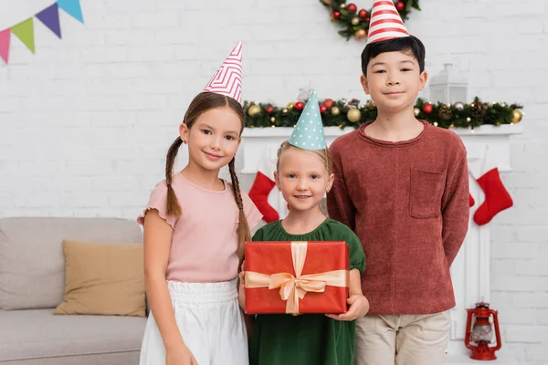 Niños multiétnicos en gorras de fiesta de pie cerca de un amigo con caja de regalo y decoración de Navidad en casa - foto de stock