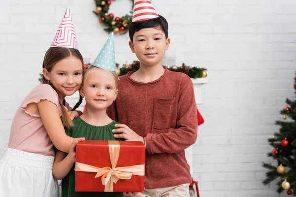 Межрасовые дети в шапочках для вечеринок обнимают друга с подарком на день рождения дома — стоковое фото
