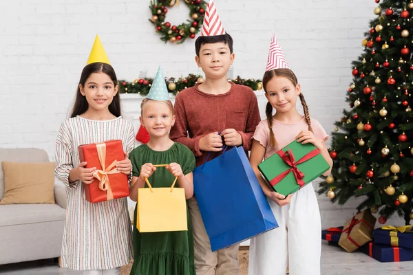 Niños multiétnicos en gorras de fiesta sosteniendo bolsas de compras y regalos cerca de la decoración borrosa de Navidad en casa - foto de stock