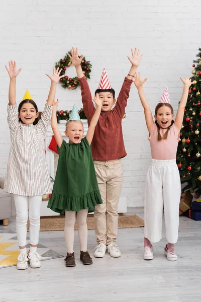 Cheerful interracial kids in party caps celebrating birthday party near blurred christmas decor at home — Stock Photo
