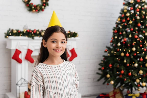 Chica sonriente en gorra de fiesta mirando a la cámara cerca del árbol de navidad arrugado en casa - foto de stock