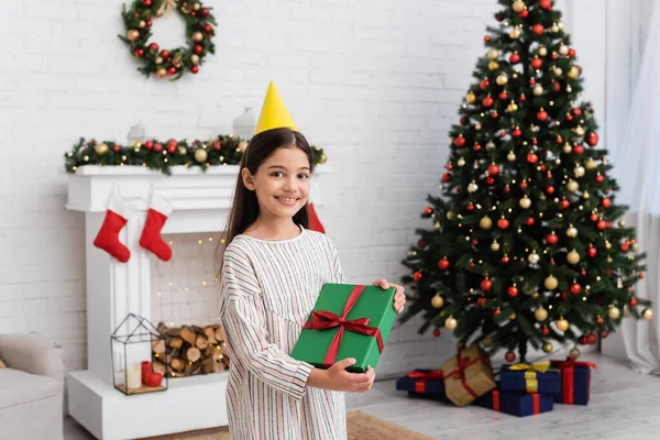 Glückliches Mädchen in Partymütze hält Geschenkbox in der Nähe verschwommenen Weihnachtsbaum und Kamin zu Hause — Stockfoto