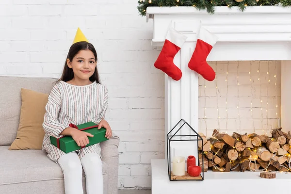 Smiling girl in party cap holding present on couch near fireplace with christmas decor at home — Stock Photo