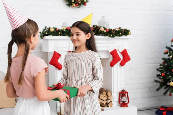 Smiling girl in party cap giving present to friend during birthday party in winter — Stock Photo