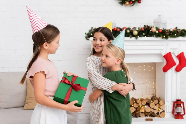 Smiling girls in party caps hugging near friend with present during birthday celebration at home — Stock Photo