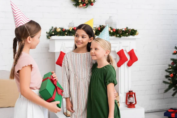 Positive girls in party caps looking at friend with gift near blurred christmas decor at home — Stock Photo