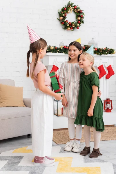 Девушки в шапочках для вечеринок смотрят на друга с подарочной коробкой возле размытых рождественских чулок дома — Stock Photo