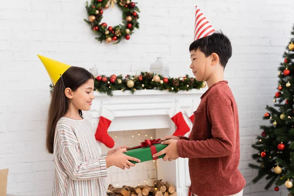 Asiático chico en partido gorra celebración regalo caja cerca sonriente amigo y Navidad decoración en casa - foto de stock