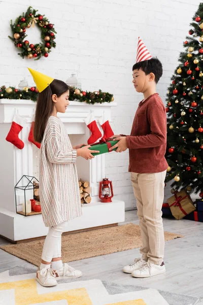 Multiethnic children in party caps holding present near christmas decor on fireplace at home — Stock Photo