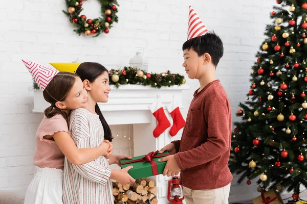 Asian boy in party cap giving present to cheerful friend near christmas decor on fireplace at home — Stock Photo