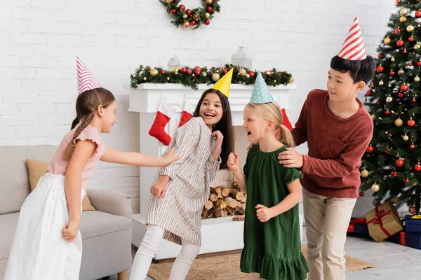 Alegre interracial niños en partido caps jugando etiqueta durante cumpleaños fiesta cerca de Navidad decoración en casa - foto de stock