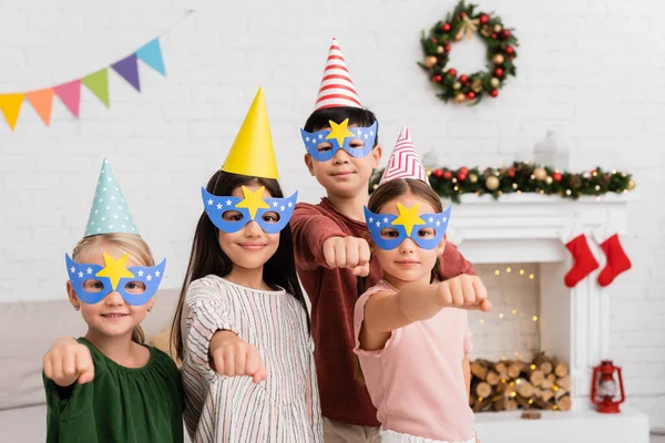 Multiethnic kids in party masks and caps gesturing at camera at home — Stock Photo