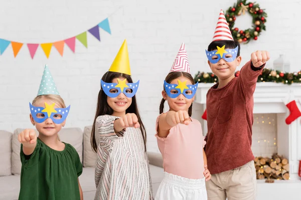 Веселі міжрасові діти в вечірніх кепках і масках жестикулюють під час вечірки на день народження вдома — стокове фото