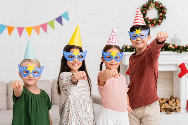 Cheerful interracial kids in party caps and masks gesturing during birthday party at home — Stock Photo
