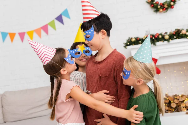 Улыбающиеся дети обнимают азиатского друга в кепке и маске во время празднования дня рождения дома — Stock Photo