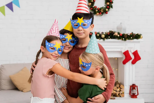Cheerful interracial children in party caps and masks hugging during birthday celebration at home — Stock Photo