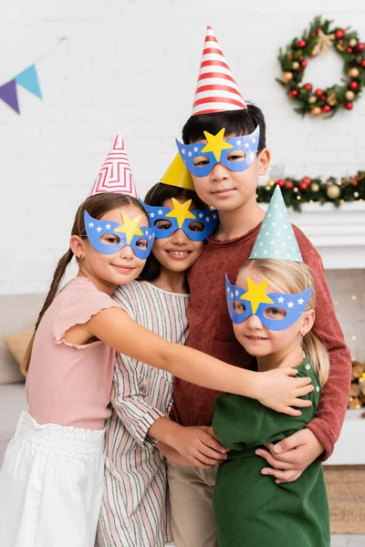 Позитивные межрасовые дети в шапочках для вечеринок и масках обнимаются и смотрят в камеру дома — стоковое фото